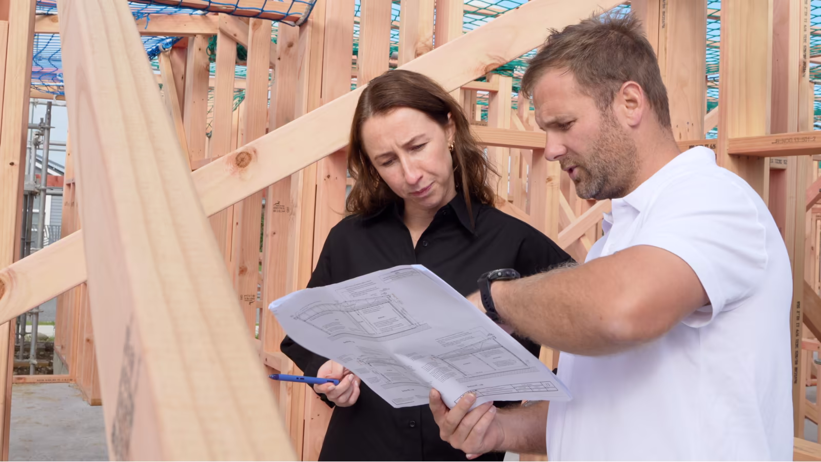 A builder and an estimator reviewing a building plan together inside a partly framed timber house on site